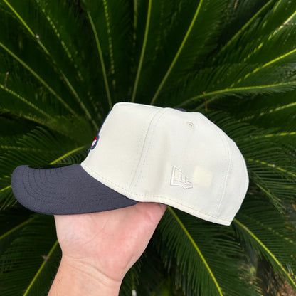 Gorra New Era beige con visera negra sostenida a mano sobre fondo de hojas verdes
