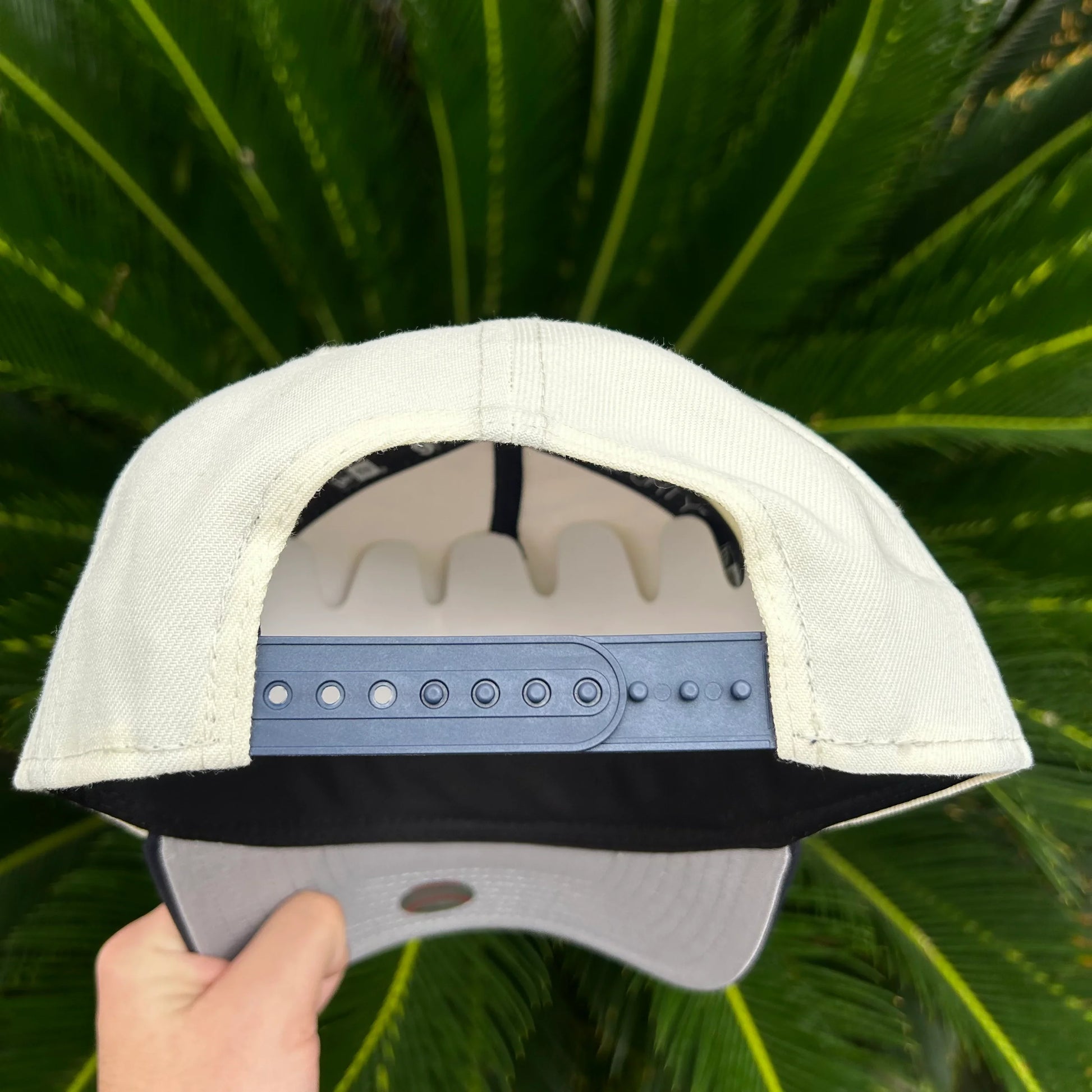 Gorra beige con cierre ajustable plástico negro y visera gris, fondo de hojas verdes