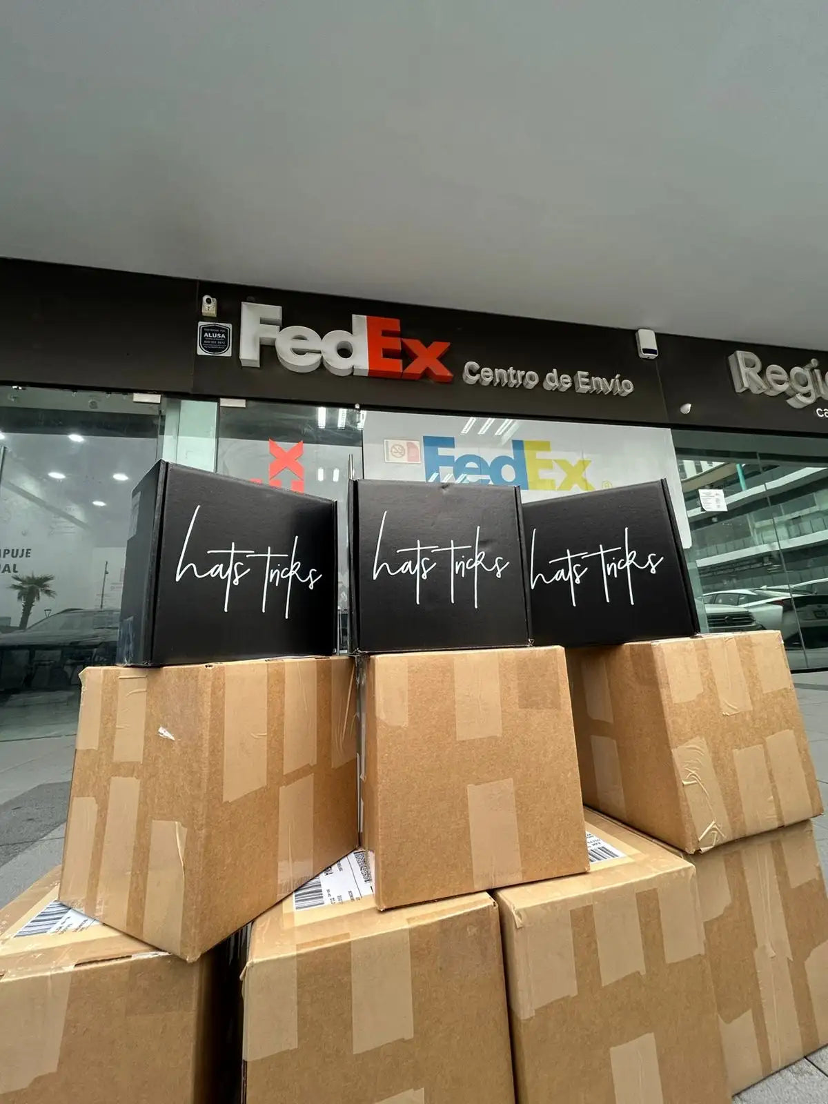 Cajas rectangulares negras con la marca 'hats tcks' en cursiva blanca, apiladas sobre cajones de envío de cartón marrón fuera de una instalación de FedEx.