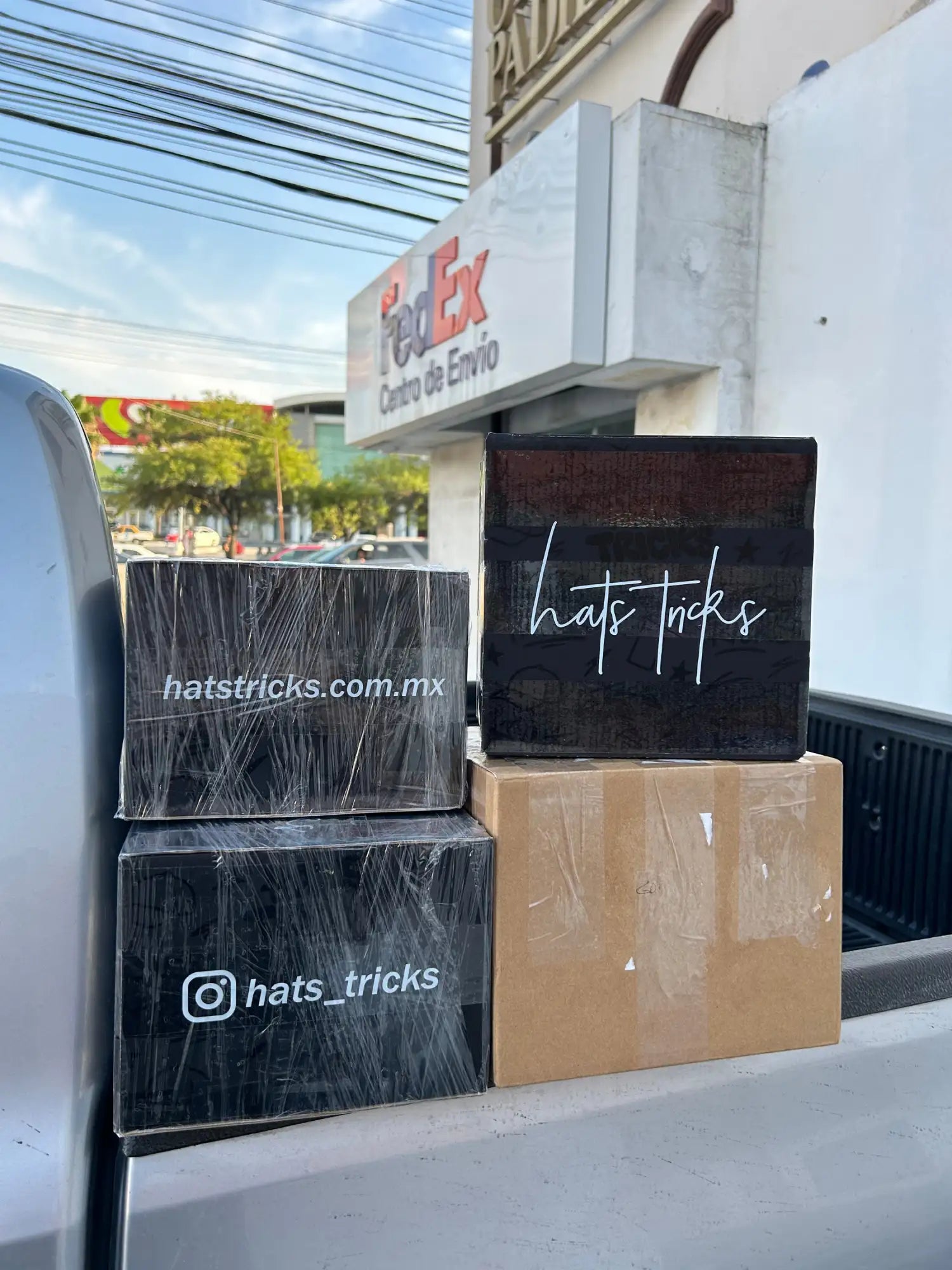 Cajas rectangulares negras con marca 'hats tricks' en cursiva blanca y una URL de sitio web, envueltas en plástico transparente.