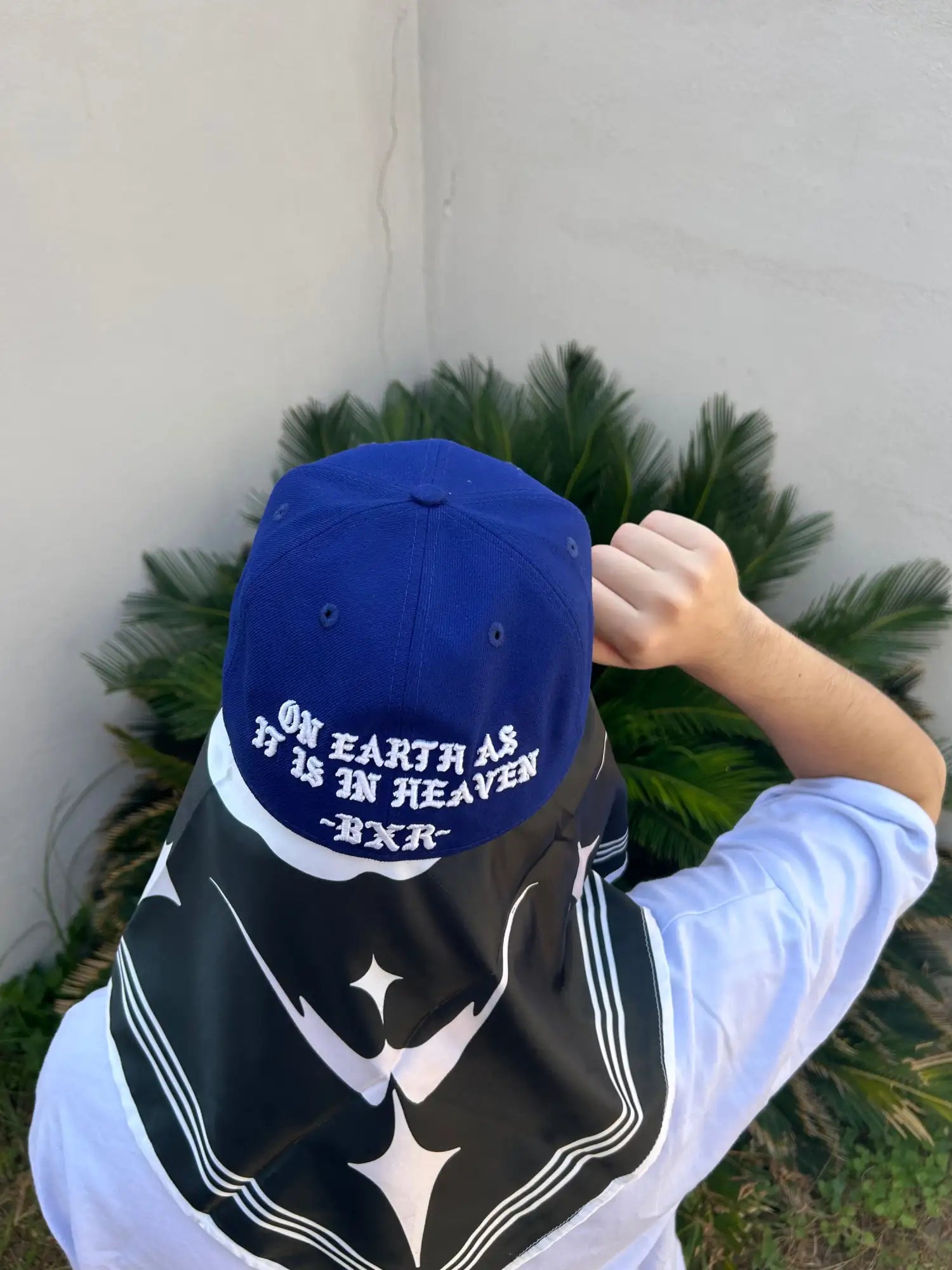 Gorra de béisbol azul con texto bordado en blanco que dice 'nuestra tierra como está en el cielo' y un pequeño '-bxr-' debajo, con una bufanda de cuello estilo marinero en blanco y negro.